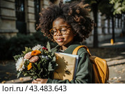 A pretty black African American woman in glasses with a bouquet of autumn flowers goes to school for the first time. A child with a school bag, a book and flowers. A child on a city street. Back to school. Стоковое AI-изображение, AI-художник Photojuli / Фотобанк Лори