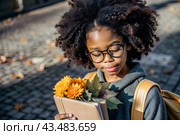 A pretty black African American woman in glasses with a bouquet of autumn flowers goes to school for the first time. A child with a school bag, a book and flowers. A child on a city street. Back to school. Стоковое AI-изображение, AI-художник Photojuli / Фотобанк Лори