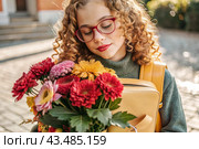 A cute little curly-haired girl with glasses and a bouquet of autumn flowers goes to school for the first time. A child with a school bag, a book and flowers. A child on a city street. Back to school. Стоковое AI-изображение, AI-художник Photojuli / Фотобанк Лори