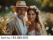 An elderly man and a young woman standing together against the backdrop of a flowering field. A man with gray hair and a beard, wearing a light linen jacket and hat, is holding a guitar. The woman, probably the bride, with delicate flowers in her hair and an elegant wedding dress, smiles. The scene is filled with warmth and joy, illuminated by soft sunlight. Стоковое AI-изображение, AI-художник Анна Кривицкая / Фотобанк Лори