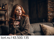 Close-up. A modern, stylish, beautiful woman with red hair and a glass of red wine in an armchair in the living room of the house. Стоковое AI-изображение, AI-художник Photojuli / Фотобанк Лори