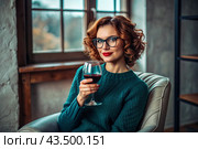 Close-up. A modern, stylish, beautiful woman with red hair and a glass of red wine in an armchair in the living room of the house. Стоковое AI-изображение, AI-художник Photojuli / Фотобанк Лори