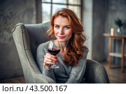 Close-up. A modern, stylish, beautiful woman with red hair and a glass of red wine in an armchair in the living room of the house. Стоковое AI-изображение, AI-художник Photojuli / Фотобанк Лори