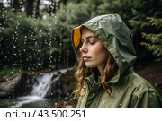 Close-up. A happy young woman in a raincoat enjoys the rain on the background of a green forest, the concept of enjoying a natural leisurely life, environment, lifestyle. Стоковое AI-изображение, AI-художник Photojuli / Фотобанк Лори