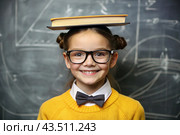 A pretty girl with glasses and books on her head. The child goes to school for the first time. A child in the classroom on the background of a blackboard. Back to school. Стоковое AI-изображение, AI-художник Photojuli / Фотобанк Лори