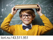 A cute boy with glasses and books on his head. The child goes to school for the first time. A child in the classroom on the background of a blackboard. Back to school. Стоковое AI-изображение, AI-художник Photojuli / Фотобанк Лори