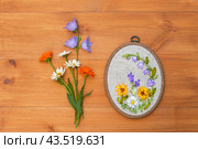 Top view of floral hand embroidery with satin ribbons of summer flowers in hoop. Bouquet of daisies, marigolds and blue bells on wooden background. Crafts and embroidery as hobby. Flat lay, copy space. Стоковое фото, фотограф Виктория Катьянова / Фотобанк Лори