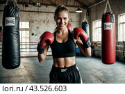 Perfect smiling female boxer is standing in ring at punching bag in an old gym, her hands in Boxing gloves. Woman workout in box training. Healthy lifestyle concept, sports in gym. Copy ad text space. Стоковое AI-изображение, AI-художник Aleksandr Golubev / Фотобанк Лори