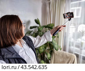Middle aged woman takes a selfie with a selfie stick in her home. Mature senior female blogger doing stream. Стоковое фото, фотограф Кривошеина Елена Леонидовна / Фотобанк Лори
