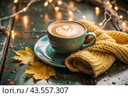 A seasonal drink. Delicious hot cappuccino in a mug with a saucer and a knitted snood, a scarf on a wooden table in the interior of the cafe. It's a rainy autumn day. Стоковое AI-изображение, AI-художник Photojuli / Фотобанк Лори