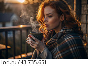 A red-haired, beautiful woman wrapped in a plaid with a mug of hot coffee on a beautiful patio in the rays of the setting sun. The girl inhales the aroma of the drink. Comfort and harmony. Стоковое AI-изображение, AI-художник Photojuli / Фотобанк Лори