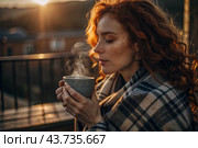 A red-haired, beautiful woman wrapped in a plaid with a mug of hot coffee on a beautiful patio in the rays of the setting sun. The girl inhales the aroma of the drink. Comfort and harmony. Стоковое AI-изображение, AI-художник Photojuli / Фотобанк Лори