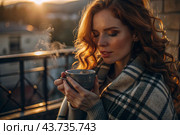 A red-haired, beautiful woman wrapped in a plaid with a mug of hot coffee on a beautiful patio in the rays of the setting sun. The girl inhales the aroma of the drink. Comfort and harmony. Стоковое AI-изображение, AI-художник Photojuli / Фотобанк Лори