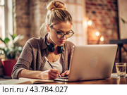 A young female freelance businessman in glasses and headphones is working on a laptop or shopping in an online store. The woman is writing something in a notebook. Стоковое AI-изображение, AI-художник Photojuli / Фотобанк Лори