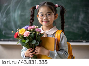 A first-grader Asian girl with glasses at the blackboard. The girl has flowers in her hands. The child goes to school for the first time. A child with a backpack, a book and flowers. Back to school. Стоковое AI-изображение, AI-художник Photojuli / Фотобанк Лори