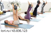Focused fit young girl practicing Pilates with diverse group, lying on mat and performing controlled circles with straight legs while holding exercise ball between ankles in sunlit fitness studio. Стоковое видео, видеограф Яков Филимонов / Фотобанк Лори
