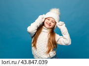 Child girl in a white sweatshirt and knitted hat with pompom on a blue background. The concept of children's emotions, joy and delight. Advertising clothing and sales. Ski resort vacation, copy space. Стоковое фото, фотограф Ирина Михайличенко / Фотобанк Лори