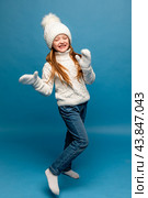 Child girl in a white sweatshirt and knitted hat with pompom on a blue background. The concept of children's emotions, joy and delight. Advertising clothing and sales. Ski resort vacation, copy space. Стоковое фото, фотограф Ирина Михайличенко / Фотобанк Лори