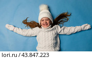 Top view child girl in a white sweatshirt and knitted hat with pompom on a blue background. children emotions, joy and delight. Advertising clothing and sales. Ski resort vacation, copy space. Стоковое фото, фотограф Ирина Михайличенко / Фотобанк Лори