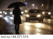 Night silhouette of woman with an umbrella on edge of busy road. Стоковое AI-изображение, AI-художник Владимир Григорьев / Фотобанк Лори