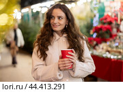 Smiling woman walking through Christmas street market with warm beverage in paper cup. Стоковое фото, фотограф Яков Филимонов / Фотобанк Лори