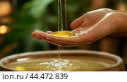 A person gracefully pours golden liquid oil from their hand into a wooden bowl during a serene moment of relaxation at a spa. Стоковое AI-изображение, AI-художник Сергей Толмачёв / Фотобанк Лори