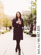 Beautiful serious smart brunette girl holding cup of coffee in hands goes walking down street of St. Petersburg in city center. Charming thoughtful woman with long dark hair wanders alone. Стоковое фото, фотограф Natalia Gdovskaia / Фотобанк Лори