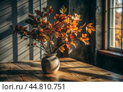 Branches with golden autumn leaves in a ceramic vase on a wooden table by the window. The concept of home comfort. Autumn decor for Halloween. Стоковое AI-изображение, AI-художник Photojuli / Фотобанк Лори