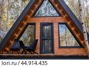 A-shaped frame house in Scandinavian style. The landscape. Quiet rest. The concept of glamping and chalet rentals. A wooden house with a veranda in nature around an autumn birch forest. Стоковое фото, фотограф Photojuli / Фотобанк Лори