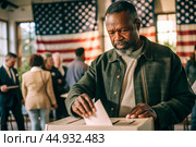 President elections. A black man votes for the new president of the United States of America and puts a form in the ballot box against the background of the American flag. Стоковое AI-изображение, AI-художник Photojuli / Фотобанк Лори