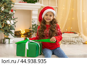 Christmas spirit. Happy little smiling girl opening gift box sitting on the floor near the Christmas tree. Happy girl unwrapping christmas present looking in box. Holidays and celebrations concept. Стоковое фото, фотограф Ирина Михайличенко / Фотобанк Лори