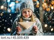 A little girl in winter clothes holding an envelope outside in snowy weather against the background of lights. A letter for Santa Claus. The winter atmosphere of a festive invitation. Стоковое AI-изображение, AI-художник Photojuli / Фотобанк Лори
