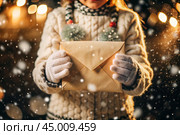 A little girl in winter clothes holding an envelope outside in snowy weather against the background of lights. A letter for Santa Claus. The winter atmosphere of a festive invitation. Стоковое AI-изображение, AI-художник Photojuli / Фотобанк Лори