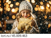 A little girl in winter clothes holding an envelope outside in snowy weather against the background of lights. A letter for Santa Claus. The winter atmosphere of a festive invitation. Стоковое AI-изображение, AI-художник Photojuli / Фотобанк Лори