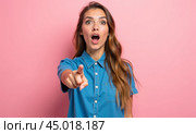 Surprised young woman in a blue shirt points directly at camera, her mouth open in amazement. Standing against pink background, excitement, shock, or unexpected discovery, marketing. Стоковое AI-изображение, AI-художник Павел Муравьев / Фотобанк Лори