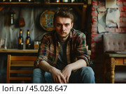 Young man wearing plaid shirt sitting and waiting in pub. Стоковое AI-изображение, AI-художник Иван Рябоконь / Фотобанк Лори