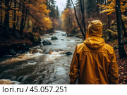 A man in a yellow rubber raincoat on the background of a huge river in a forest in the mountains. The guy travels and enjoys life and the environment. Tourism. Стоковое AI-изображение, AI-художник Photojuli / Фотобанк Лори