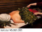 Russian bathhouse. Man in bath hat lying and sweats on wooden bench in sauna with hot steam. Hands with broom whip steamed body in spa complex. Wellness, self-care, healthy concept. Copy ad text space. Стоковое фото, фотограф Aleksandr Golubev / Фотобанк Лори