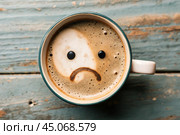 A mug of coffee with a sad smiley face on the foam on the blue table. Top view, flat lay. Стоковое AI-изображение, AI-художник Photojuli / Фотобанк Лори