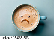 A mug of coffee with a sad smiley face on the foam on the blue table. Top view, flat lay. Стоковое AI-изображение, AI-художник Photojuli / Фотобанк Лори