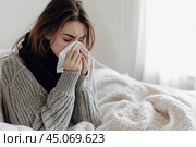 Young sick woman with cold or flue sitting on grey couch, holding a tissue to clear, blowing runny nose. Sneezing Girl under gray plaid blanket with handkerchief to dripping nose, blowing snot at home. Стоковое AI-изображение, AI-художник Марина Демидюк / Фотобанк Лори