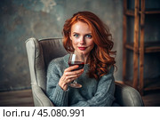 Close-up. A modern, stylish, beautiful woman with red hair and a glass of red wine in an armchair in the living room of the house. Стоковое AI-изображение, AI-художник Photojuli / Фотобанк Лори