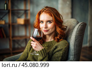 Close-up. A modern, stylish, beautiful woman with red hair and a glass of red wine in an armchair in the living room of the house. Стоковое AI-изображение, AI-художник Photojuli / Фотобанк Лори