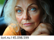 Close-up of an elderly woman's face with grey hair and bright blue eyes. She looks straight into the camera, her face conveying calm and deep thought. Her skin has pronounced wrinkles, which emphasizes her age and life experience. The light falls softly on her face, giving the photo a natural and warm feel. The woman's hands rest relaxedly on her chin, creating an atmosphere of trust and openness. Редакционное фото, фотограф Анна Кривицкая / Фотобанк Лори