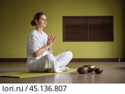 Woman practicing Kundalini yoga, sitting in comfortable position on mat and doing lotus mudra from loneliness, practicing in white clothes in room with meditation bowls placed nearby. Стоковое фото, фотограф Сергей Чайко / Фотобанк Лори