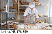 Smiling confident professional female baker wearing striped apron holding wooden rolling pin, standing near floured worktable in bakery, surrounded by freshly baked bread and baking tools. Стоковое видео, видеограф Яков Филимонов / Фотобанк Лори