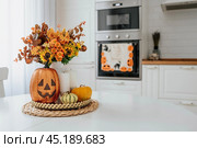 A vase of flowers,a jack pumpkin and candles on a tray. In the background - the interior of a white kitchen in Scandi style. The concept of home and comfort. Autumn decor for the Halloween holiday. Стоковое фото, фотограф Photojuli / Фотобанк Лори