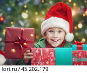 Smiling boy in red Santa Claus Christmas hat with gifts near Christmas tree. Стоковое AI-изображение, AI-художник Сергей Елизарьев / Фотобанк Лори