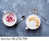 Two greek yogurt with jam and banana in white bowl on grey blue concrete stone table, top view. Стоковое фото, фотограф Natalia Gdovskaia / Фотобанк Лори