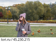 On a warm autumn day, a woman walks through the park. The girl is drinking coffee and talking on the phone. Стоковое фото, фотограф Photojuli / Фотобанк Лори